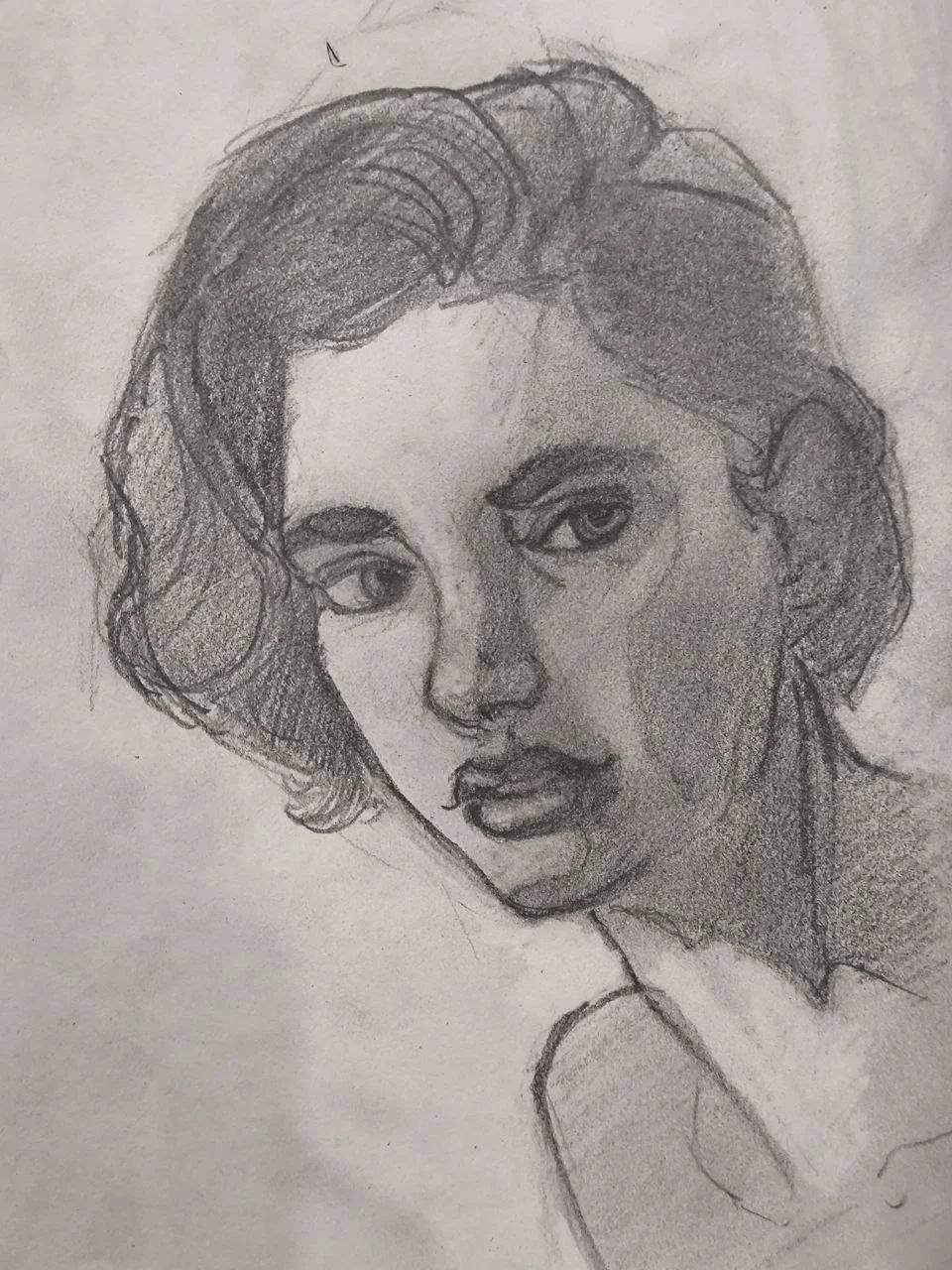Graphite portrait sketch of a person with short, wavy hair and a calm, sideways gaze; strong shading defines the nose, lips, and the shadowed side of the face, with the neck and shoulders loosely outlined on smudged paper.