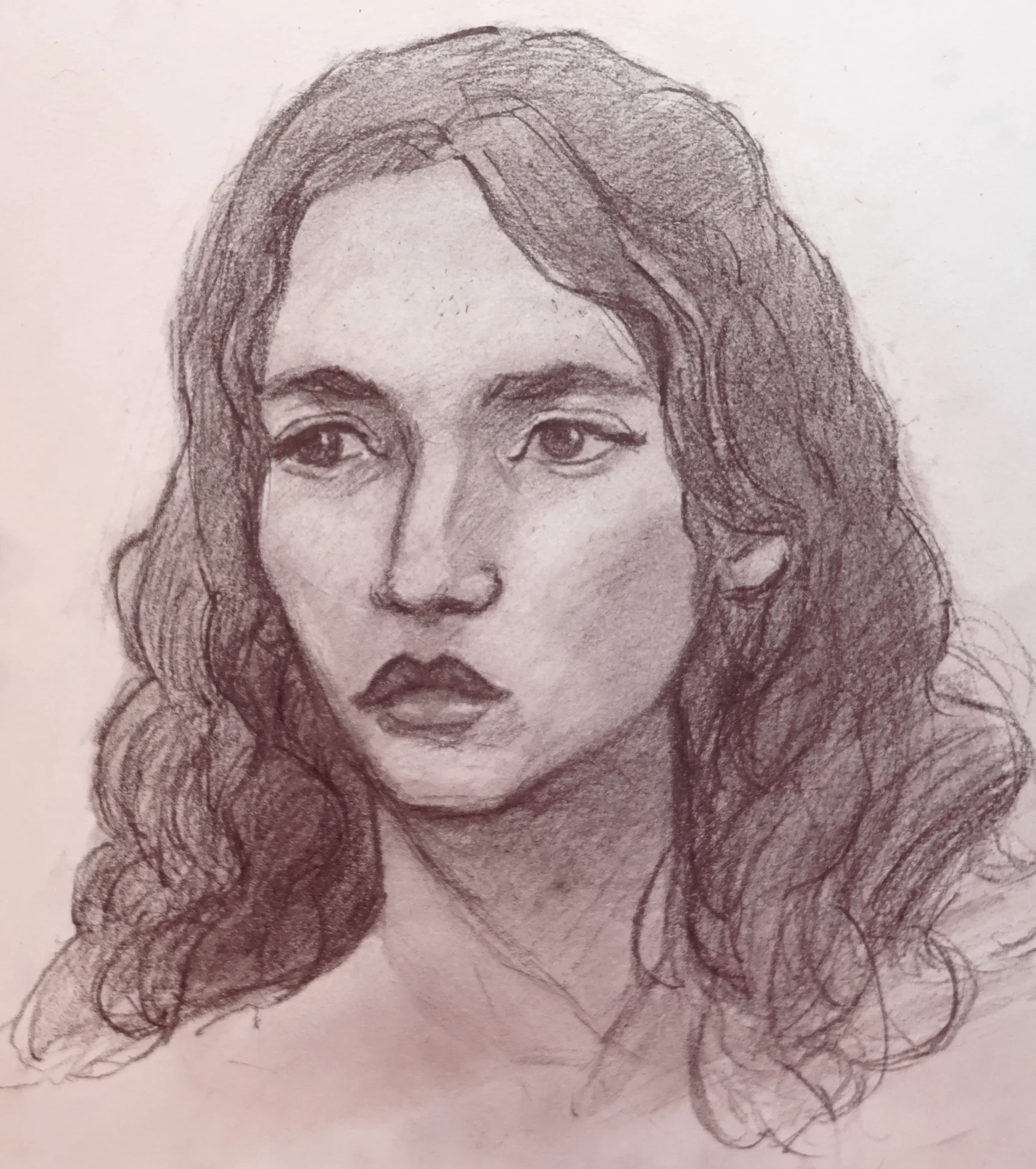 Pencil portrait drawing of a young woman with long wavy hair, looking slightly to the side. Soft shading defines the cheekbones, nose, and lips, with darker shadows under the chin and around the hair.