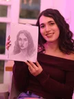 The woman from the first portrait holds up a sketchbook with her graphite portrait, smiling under soft pink light.