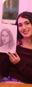 The woman from the first portrait holds up a sketchbook with her graphite portrait, smiling under soft pink light.