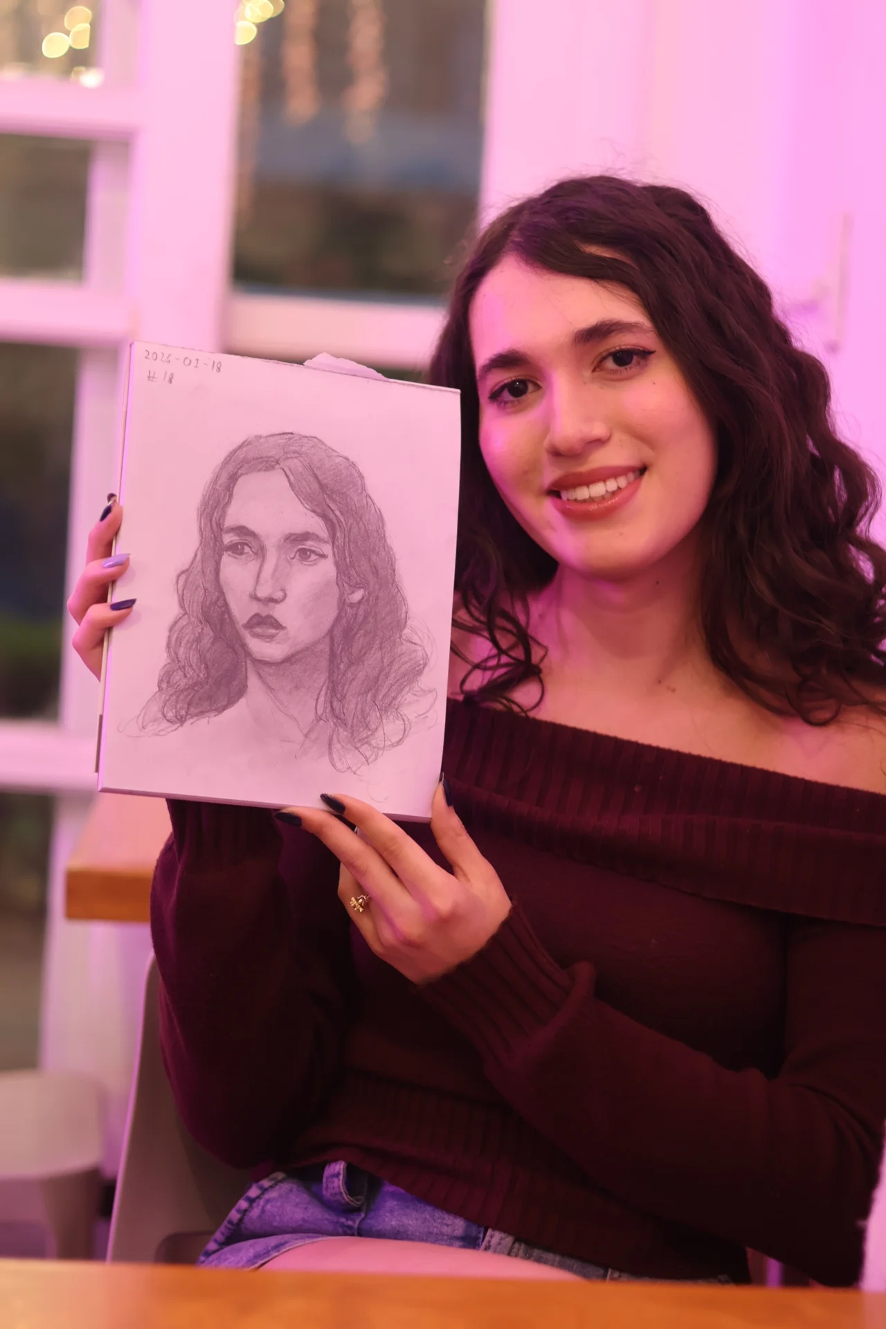 The woman from the first portrait holds up a sketchbook with her graphite portrait, smiling under soft pink light.