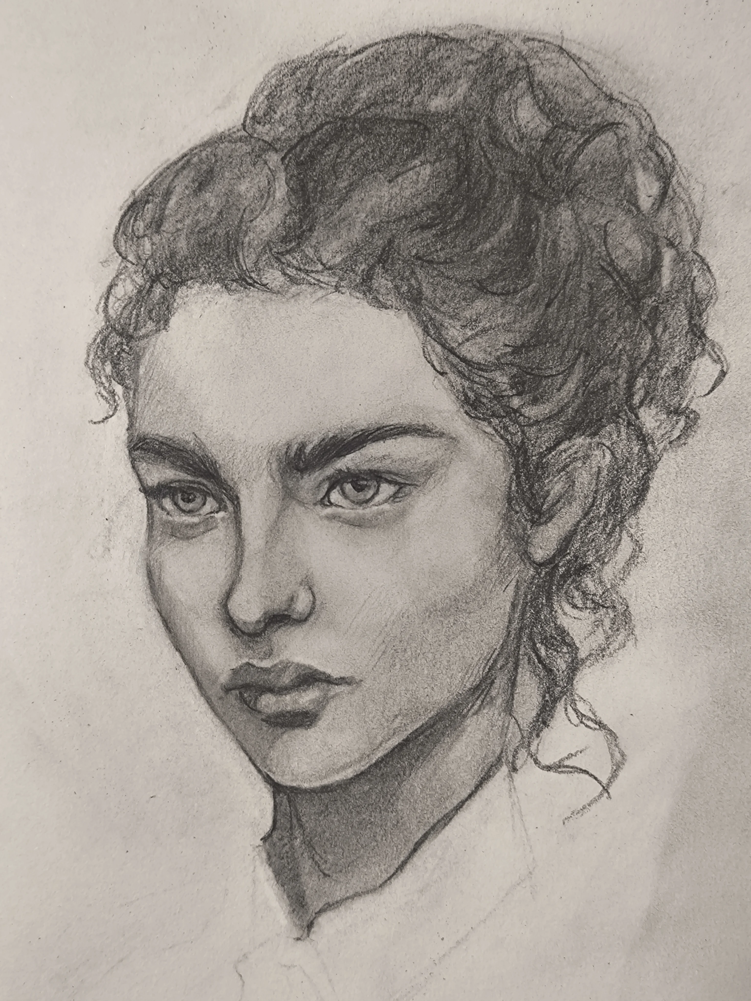 Graphite portrait sketch of a young woman in three-quarter view, looking slightly to the left. She has thick eyebrows, calm eyes, and full lips, with strong shading under the cheekbone and neck. Her curly hair is tied back with loose strands on the side.