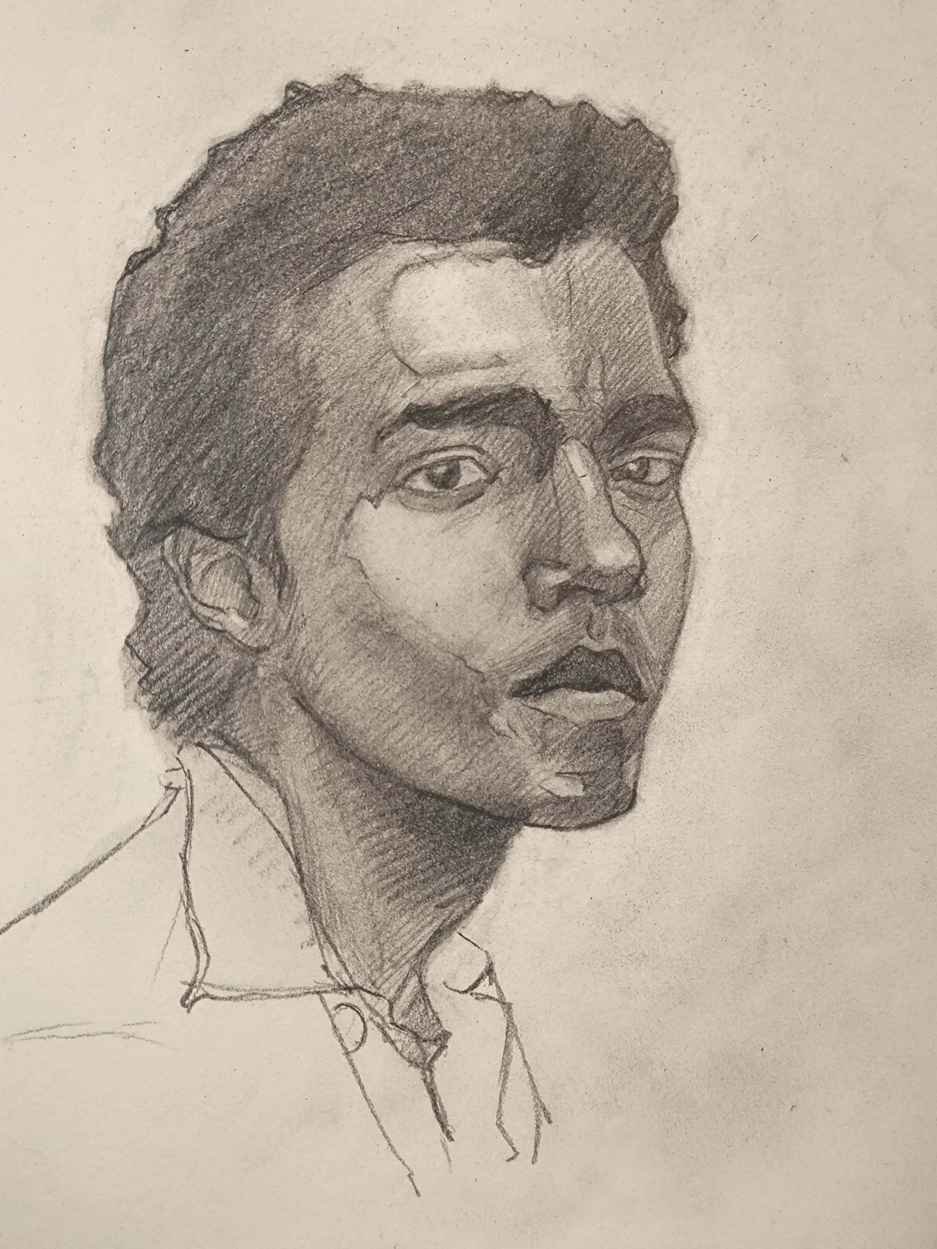 Pencil portrait sketch of a young man in 3/4 view, looking slightly to the right. He has short curly hair, strong cheekbones, and full lips. The face is shaded with soft graphite tones and crosshatching, and there is a loose outline of a shirt collar.
