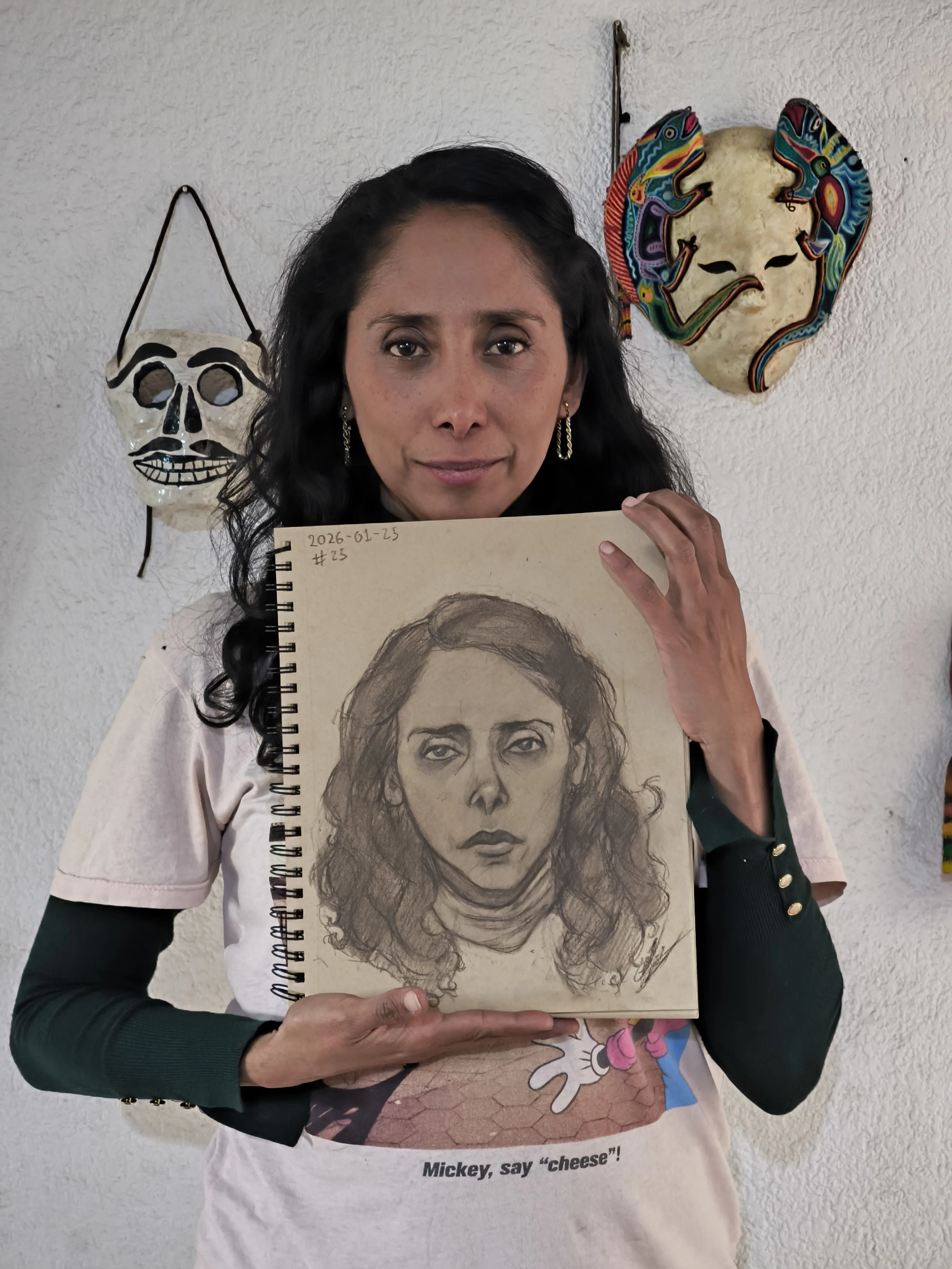 A woman with long dark hair stands against a white textured wall, holding a spiral sketchbook with the portrait from the first picture. Two decorative masks hang on the wall behind her.