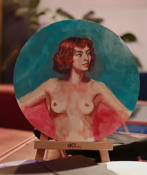 Small round painting on a stand, shows a red haired woman with bare torso, looking to the side. Soft brush strokes and warm skin tones with teal background, scene has cozy desk and notebook in front.