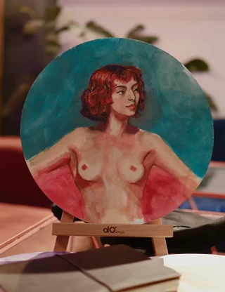 Small round painting on a stand, shows a red haired woman with bare torso, looking to the side. Soft brush strokes and warm skin tones with teal background, scene has cozy desk and notebook in front.