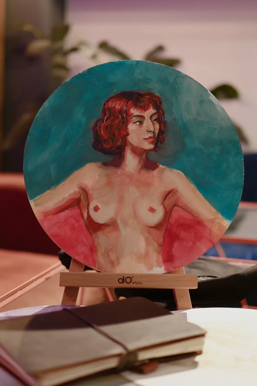 Small round painting on a stand, shows a red haired woman with bare torso, looking to the side. Soft brush strokes and warm skin tones with teal background, scene has cozy desk and notebook in front.