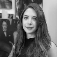 A black-and-white photograph of the artist Elena Manero. She is shown with long, straight hair and a relaxed smile, looking directly at the camera. Several oil paintings are visible in the background.